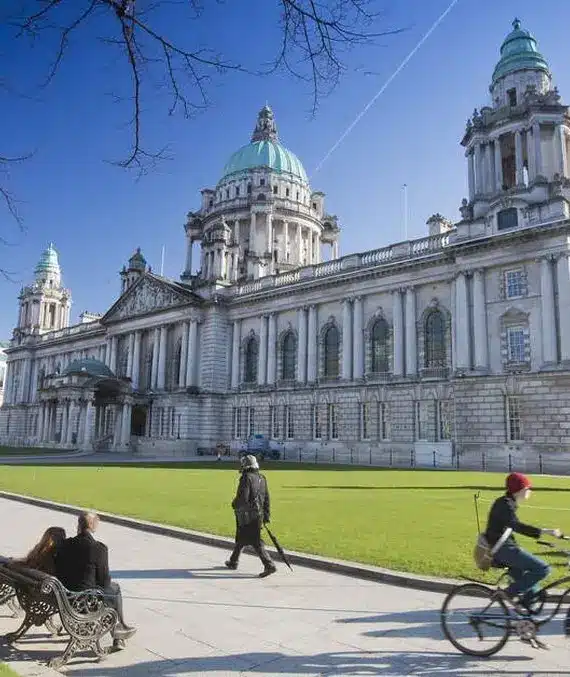 People walk and cycle past a grand historic building with domes and columns, likely a town hall, on a sunny day. The foreground features green lawns, benches, and leafless trees.
