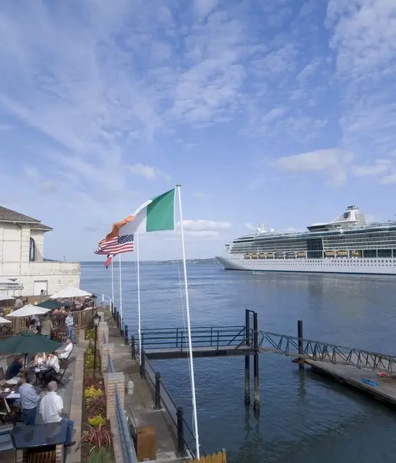 People dine on a waterfront terrace next to a quay, with Irish, American, and European Union flags flying. A large white cruise ship sails past under a blue sky with scattered clouds.