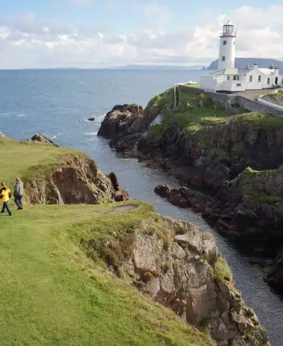 Two people walk on grassy cliffs near the sea, with a white lighthouse and its buildings perched on a rocky outcrop in the background under a partly cloudy sky.