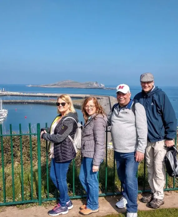Four adults standing and smiling in front of a green fence, with a marina, boats, and blue sea in the background on a sunny day. An island is visible in the distance under a clear sky.