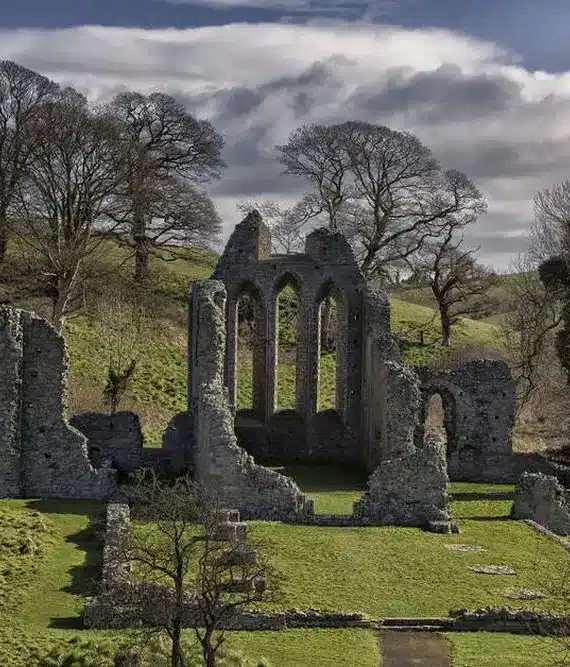 Ancient stone ruins of a gothic abbey stand on a grassy hill, surrounded by leafless trees and rolling green countryside under a cloudy sky.