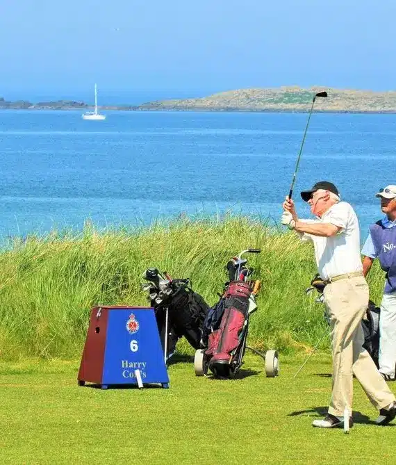 Four men stand on a grassy golf course by the sea, with one preparing to swing. Two golf bags and a sign reading “6 Harry” are nearby. The blue sea, a sailing boat, and distant land are visible in the background.