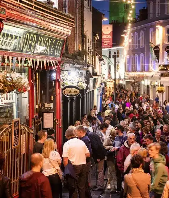 A lively crowd fills a narrow, brightly lit street lined with pubs and hanging flower baskets at night, creating a festive and bustling atmosphere.
