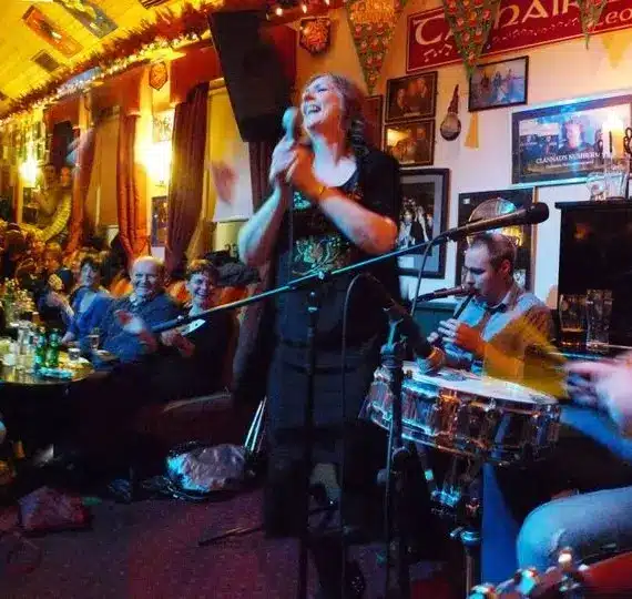 A lively pub scene with a band performing on stage. The female singer stands at the microphone, accompanied by a drummer and guitarist, whilst a large, cheerful crowd watches and enjoys drinks in the cosy, decorated venue.