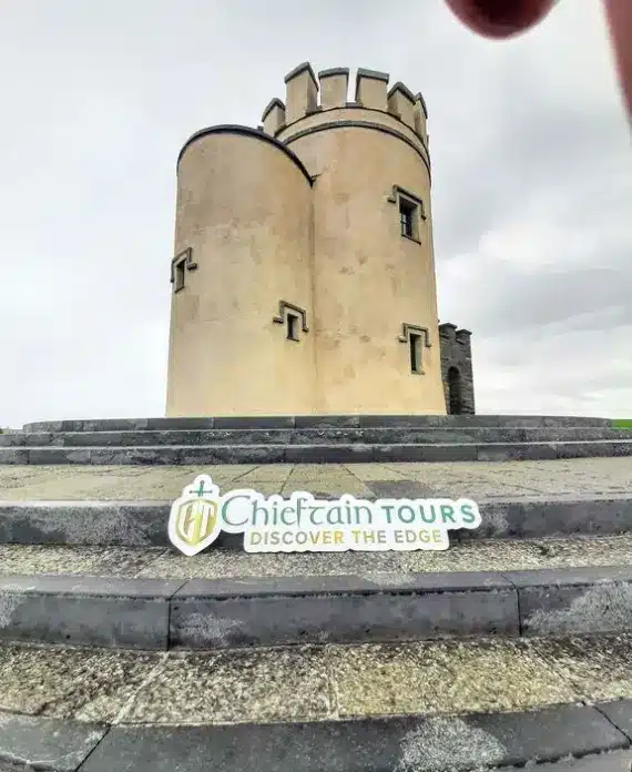 A round stone tower with crenellations stands on a raised platform with steps. A "Chieftain Tours Discover the Edge" sign is displayed at the base. The sky is cloudy and a hand partially covers the upper right corner.