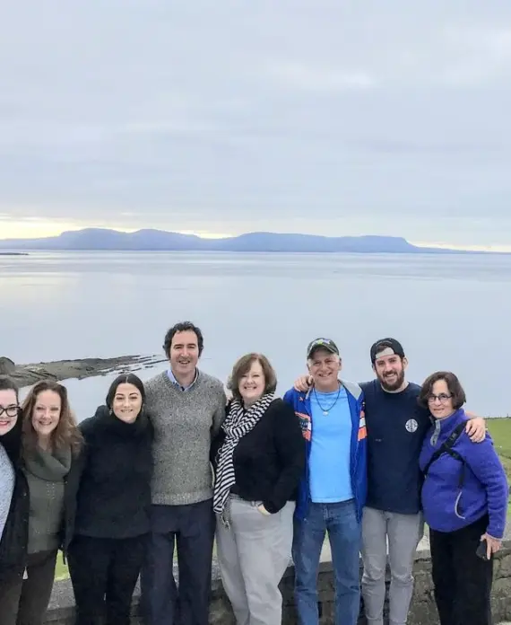 Eight people stand closely together, smiling at the camera with an expansive scenic backdrop of calm water, distant hills, and green grassy landscape under a cloudy sky. They appear to be enjoying a group outing.