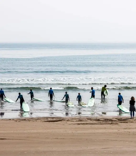 A group of people in wetsuits carrying surfboards walk towards the sea from a sandy beach, while one person stands on the shore watching them. The sea is calm with gentle waves.