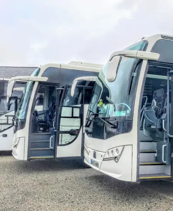 Three white coach buses are parked side by side in a car park, with their front doors open. The background includes a building with a dark roof and some greenery. The sky appears cloudy.