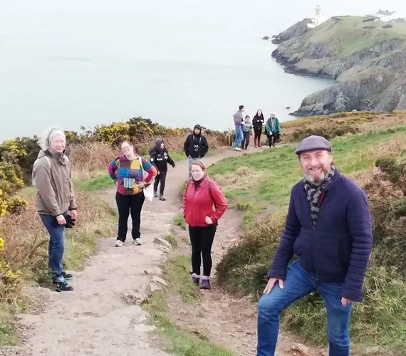 A group of people walking along a coastal cliffside path lined with green bushes and yellow flowers, with the sea and a lighthouse visible in the background under an overcast sky.
