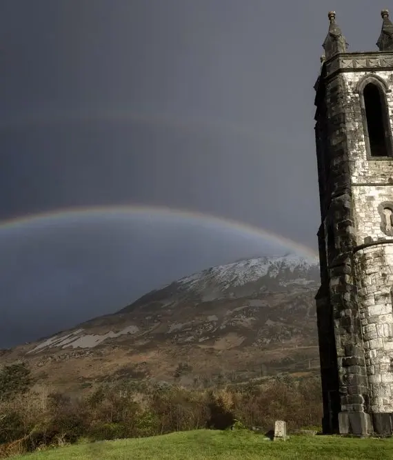 A tall stone church stands in front of a mountain under a dark sky with a double rainbow arching overhead. The landscape is green and lightly wooded, and the mountain has a dusting of snow.