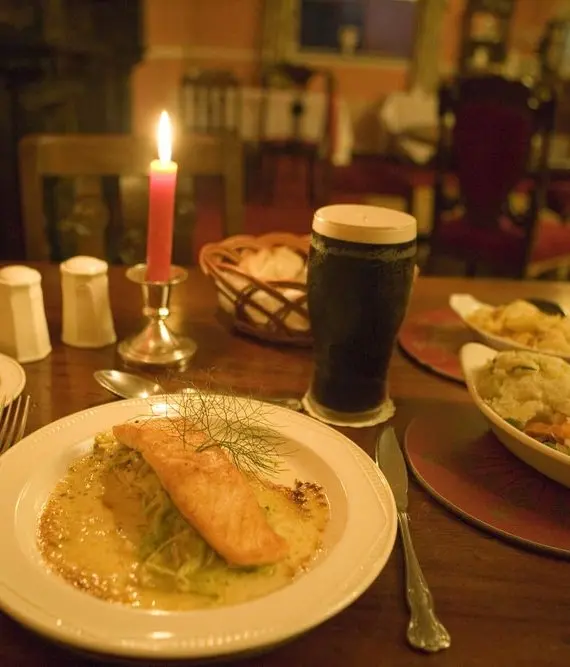 A cosy restaurant table set for one with a lit candle, a pint of dark beer, bread with butter, a plate of grilled salmon with sauce, and a side dish of mashed potatoes and mixed vegetables.