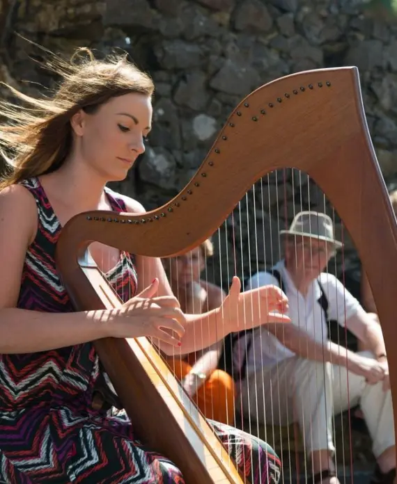 A woman in a patterned dress plays a wooden harp outdoors, with wind blowing her hair. Three people sit in the background on stone steps, watching her performance.