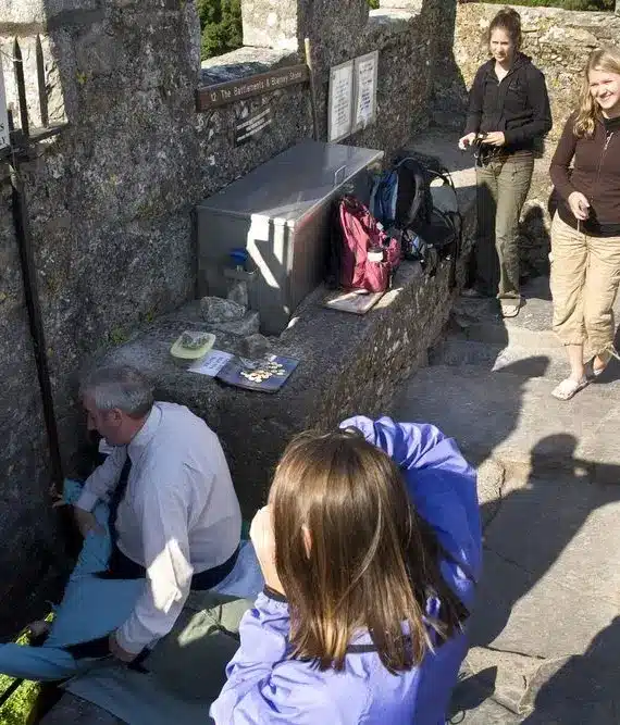 Several people gather on a stone platform outdoors. One person is preparing to lean backwards against a wall, possibly to kiss the Blarney Stone, whilst others watch and wait their turn. Rucksacks and informational signs are visible.