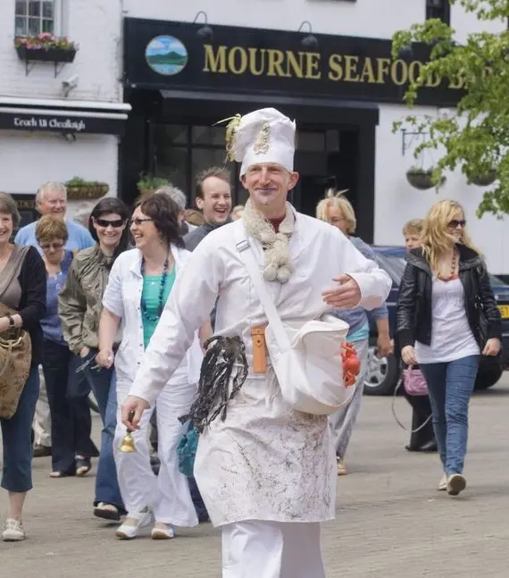 A man dressed as a chef, wearing white clothes and a hat decorated with utensils, walks smiling in front of a group of people in a town square near a seafood restaurant. The group looks cheerful and relaxed.