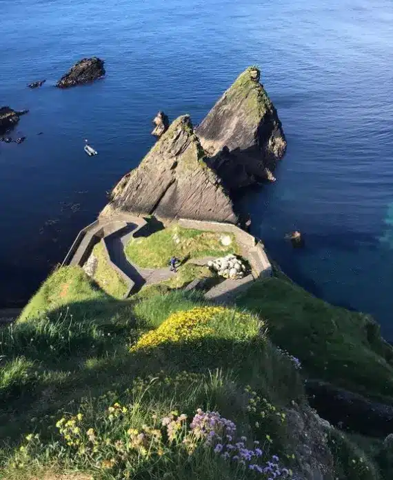 A steep, grassy cliff overlooks a winding stone path and pier beside jagged rock formations jutting out into the blue sea, with a small boat floating nearby.