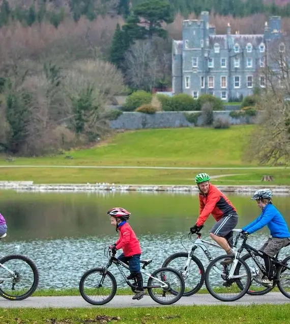 Four people, two adults and two children, ride bicycles on a path beside a lake in a park. A large, castle-like building stands in the background among trees and greenery.