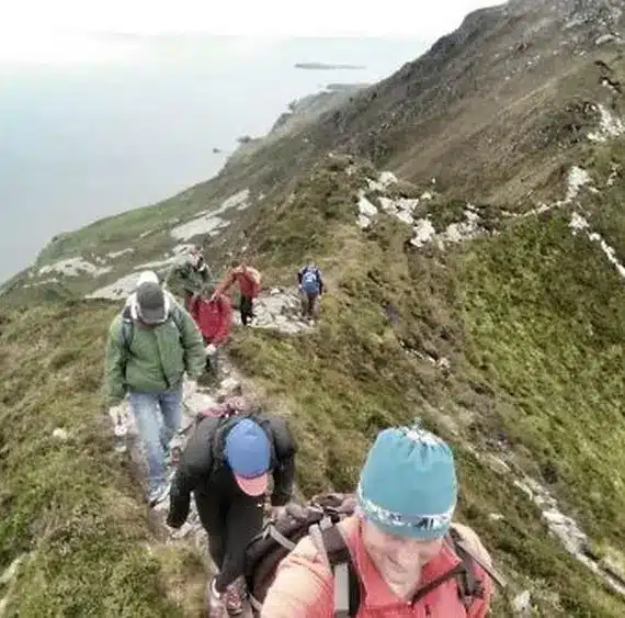 A group of hikers, wearing rucksacks and outdoor gear, walk along a narrow, grassy mountain ridge with steep slopes, overlooking the sea and coastline in the distance.