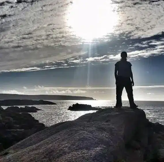 A person stands on a rocky cliff overlooking the sea, silhouetted against a dramatic sky with the bright sun shining through scattered clouds.