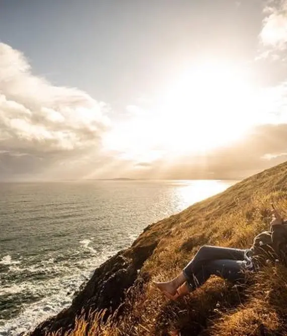 A person sits on a grassy hillside overlooking the sea at sunset, with golden sunlight streaming through clouds and illuminating the landscape. Waves break gently against the shore below.
