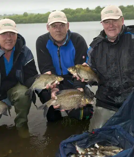 Three men wearing matching hats and waterproof jackets kneel by a lake, smiling and holding several fish in their hands. More fish are collected in a blue net on the ground at their feet. Trees and cloudy sky are in the background.