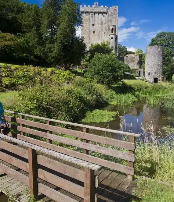 A couple walks across a wooden bridge over a stream, with lush greenery and a historic stone castle and tower in the background under a bright blue sky.