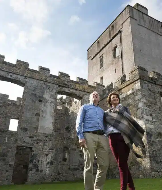 A man and a woman stand on grass in front of a large stone castle with tall towers and battlements, looking up and smiling under a partly cloudy sky.