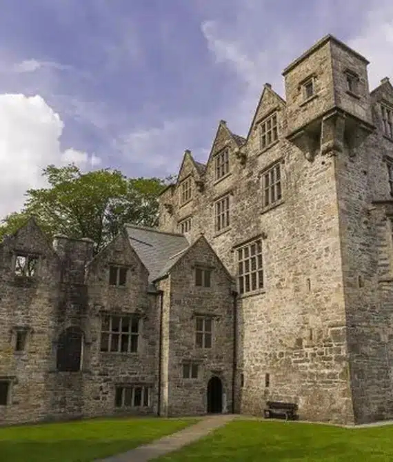 A large stone castle with tall, narrow windows and pointed gables stands on a grassy lawn under a partly cloudy sky. Trees surround the historic building.