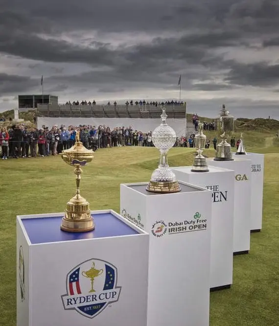 Four golf trophies, including the Ryder Cup, are displayed on stands on a golf course, with crowds and an empty grandstand in the background under a cloudy sky.