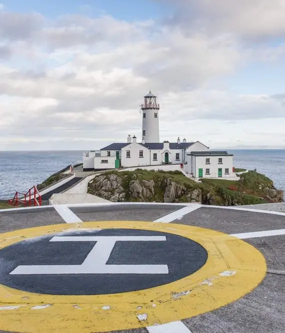 A white lighthouse with attached buildings sits on a rocky coast, surrounded by the sea. In the foreground is a large helipad marked with a yellow circle and an ‘H’. The sky is partly cloudy.