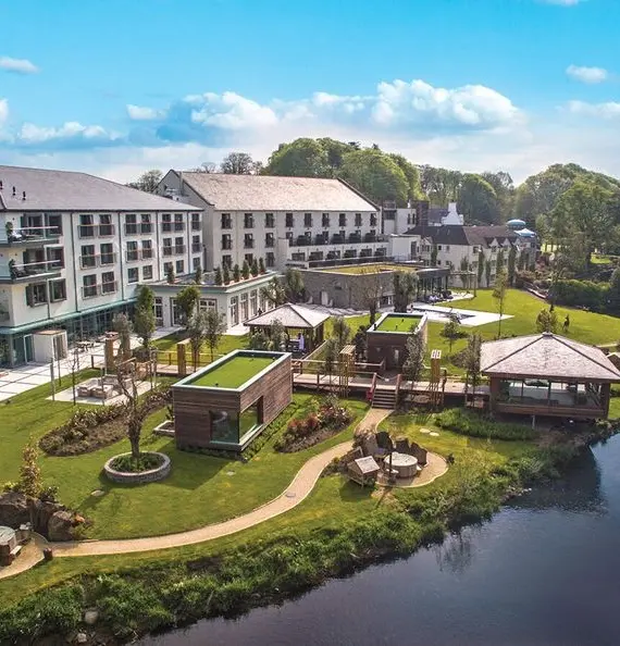 Luxurious riverside resort with modern buildings, manicured lawns, patio seating, outdoor pavilions, and a scenic river bordered by trees under a bright blue sky with fluffy white clouds.