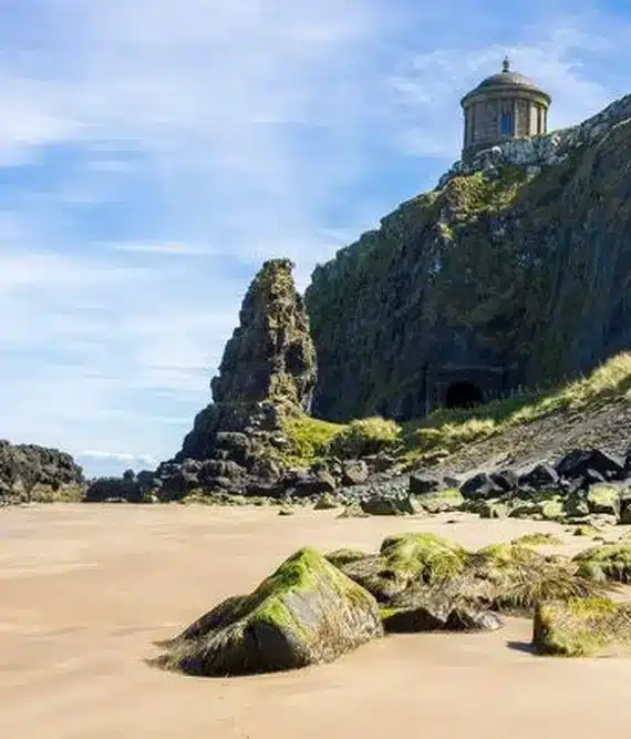 A sandy beach with scattered rocks, a steep grassy cliff, and a small circular stone building with a domed roof perched on top, under a blue sky with light clouds.