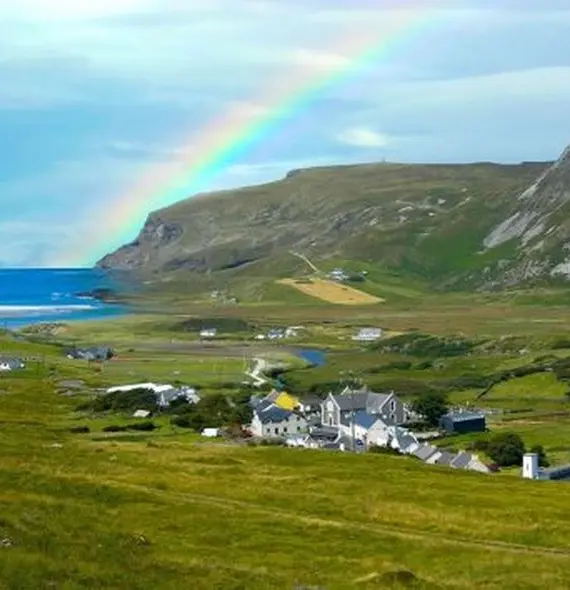 A vibrant rainbow arches over a coastal village with green hills, white houses, and cliffs, ending near the blue sea under a partly cloudy sky.