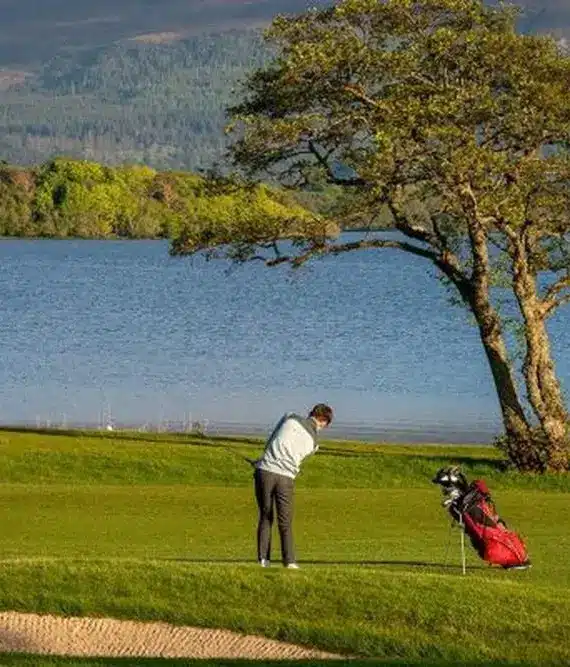A golfer stands on a green near a sand bunker, mid-swing, with a golf bag beside them. A lake and trees are in the background, beneath distant mountains.