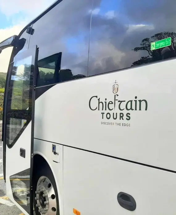 A white tour coach labelled "Chieftain Tours Discover the Edge" is parked on a road near a stone wall and greenery under a partly cloudy sky.