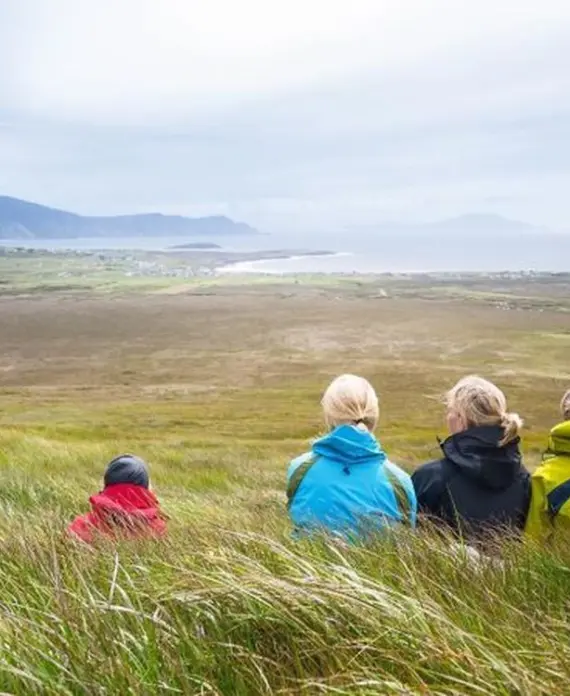 Four people wearing colourful jackets sit in tall grass, overlooking a vast, green landscape with distant hills and a cloudy sky.