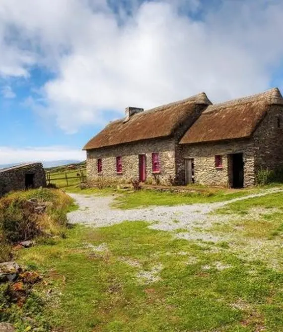 A rustic stone cottage with a thatched roof sits on a grassy landscape under a partly cloudy sky, surrounded by hills and greenery, with a distant view of the sea.