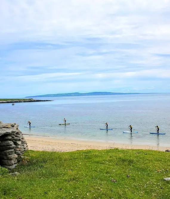 A group of people paddleboarding on calm, clear water near a sandy beach with a stone wall and grassy area in the foreground under a partly cloudy sky.