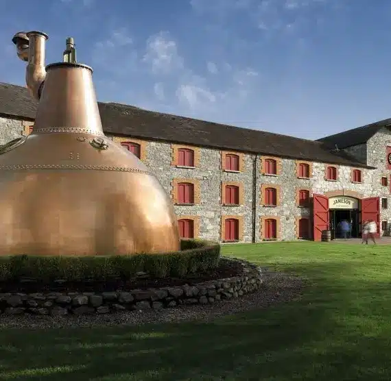 A large copper pot still is displayed on grass in front of a historic stone building with red doors and windows, under a blue sky. Several people walk near the building’s entrance.