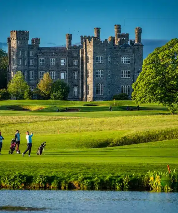Four people play golf on a green course with golf bags, set against a backdrop of a large stone castle surrounded by trees and greenery under a clear blue sky, with a pond in the foreground.