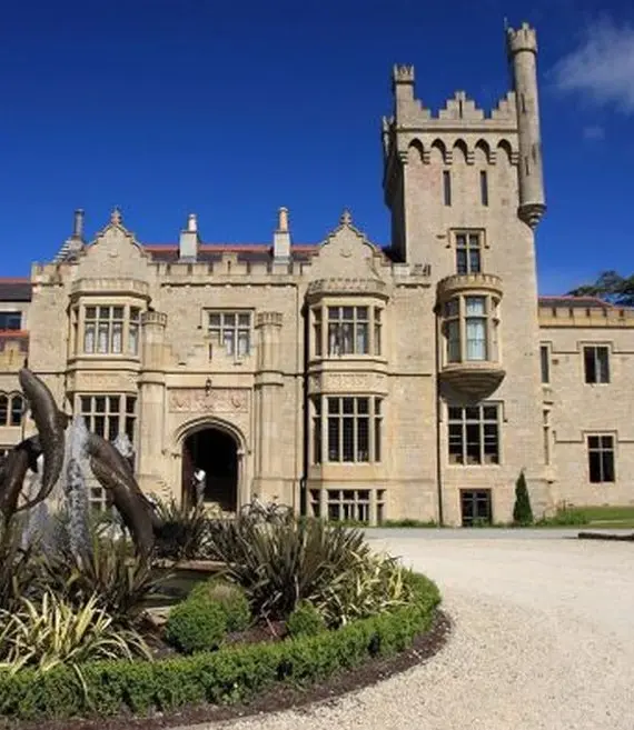 A large stone castle with Gothic-style architecture, arched windows, and a tall tower, set against a blue sky, with a roundabout garden and gravel drive in the foreground.