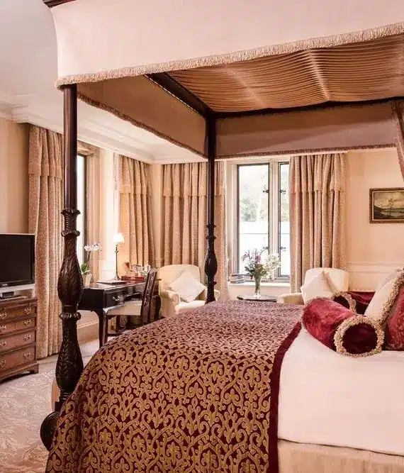Elegant hotel room with a four-poster bed, ornate bedding, classic wooden furniture, a TV on a chest of drawers, armchairs by the window, and soft beige curtains creating a luxurious, cosy atmosphere.