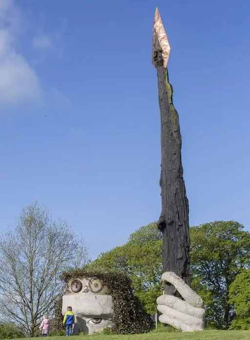 A large outdoor sculpture of a face with round eyes and wild hair, holding a tall, charred stick with an orange tip; two people stand near its base on a grassy area under a blue sky.