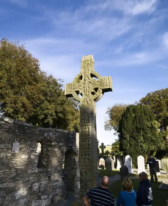 A tall, intricately carved Celtic cross stands in a graveyard, surrounded by old stone walls, trees, and several people looking up at the monument under a bright blue sky.