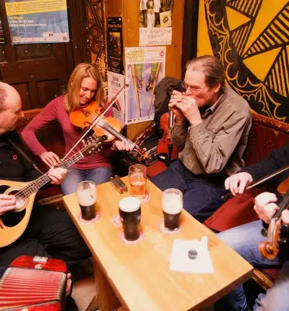 Four musicians play traditional instruments around a small wooden table with pints of dark beer in a cosy, warmly lit pub decorated with posters and art on the walls.