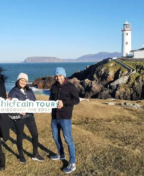 Three people stand on grassy cliffs by the sea, holding a "Chieftain Tour: Discover the Edge" sign. A white lighthouse and buildings are visible on the rocky coastline under a clear blue sky.