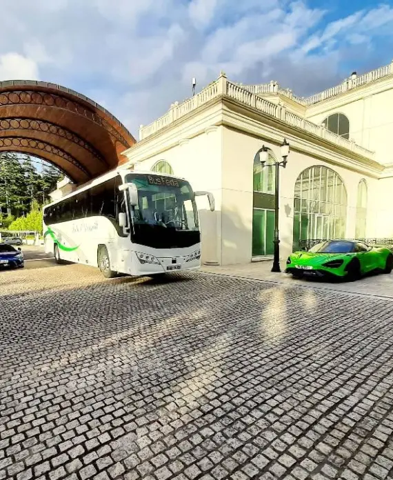 A white coach and three luxury sports cars, including a green one, are parked on a cobbled driveway next to a grand building with large arched windows and an ornate wooden canopy. Trees and blue sky are in the background.