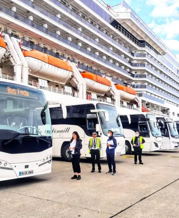 Several white tour coaches are parked next to a large cruise ship. Crew members and passengers stand on the quay, some talking, while people walk towards the coaches under a partly cloudy sky.