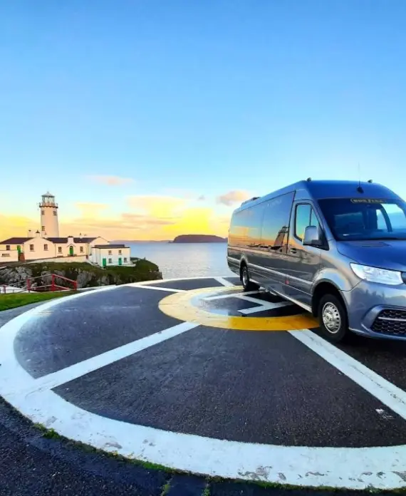 A silver Mercedes-Benz van is parked on a helipad near the coast, with a lighthouse and buildings on a small peninsula in the background, under a blue and orange sky at sunset.