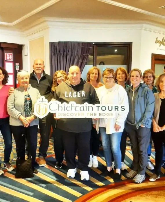 A group of 14 smiling adults pose indoors on a patterned carpet, holding a "Chieftain Tours: Discover the Edge" sign. They stand together in a hotel lobby with warm lighting and wood decor.