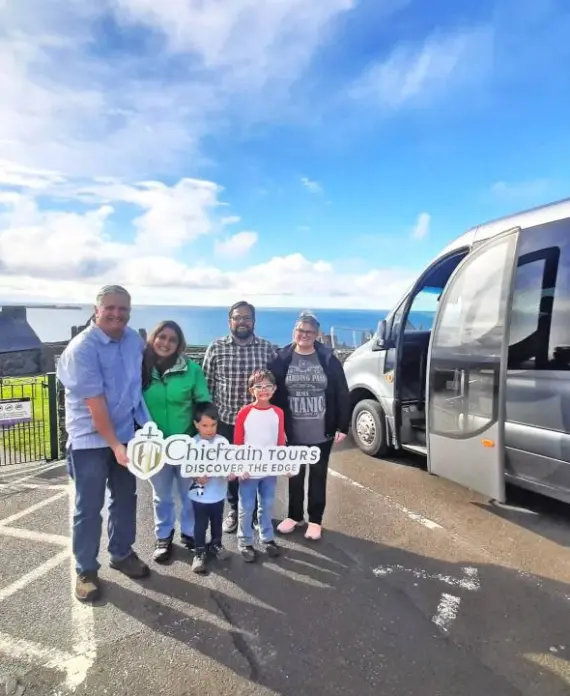 A group of six people, including children and adults, stand smiling next to a silver van in a car park. One person holds a "Chieftain Tours" sign. The background shows buildings, a fence, and a partly cloudy sky.